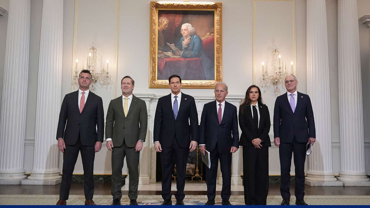 A group of six formally dressed officials standing side by side in an ornate room, with chandeliers on either side and a large framed portrait hanging above a fireplace behind them.