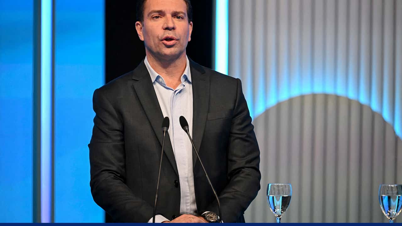 A man in a suit speaking at a podium with microphones, delivering a speech at a formal event, with a glass of water placed beside him and a blue-lit backdrop behind.