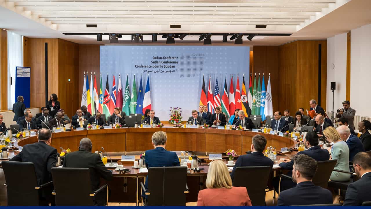 Delegates from multiple countries sit around a large circular table at an international conference on Sudan, with national flags displayed behind the panel.