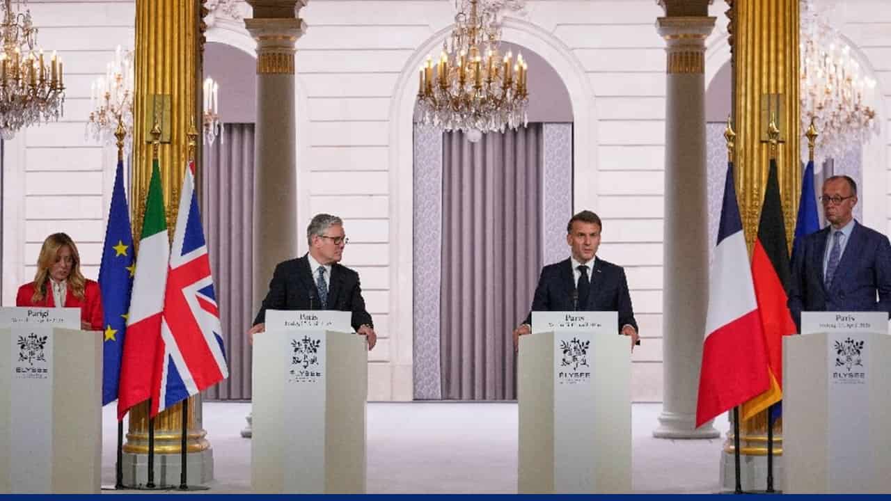 Leaders stand at podiums during a formal press conference at the Élysée Palace, with national flags of the European Union, Italy, United Kingdom, France, and Germany displayed behind them. Elegant chandeliers and gold-trimmed columns frame the setting.