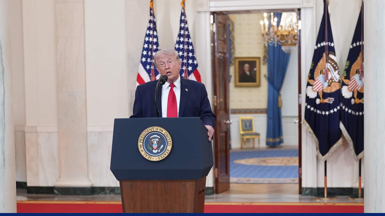 A man in a suit and red tie speaks at a podium bearing the U.S. presidential seal, with American flags behind him and a formal room visible through an open doorway.
