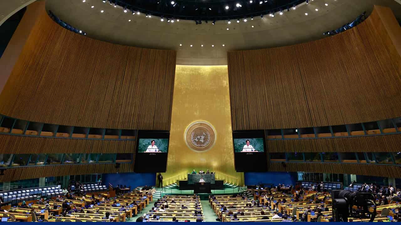 A wide view of the United Nations General Assembly hall with delegates seated in rows, a speaker at the central podium beneath a large UN emblem, and two large screens displaying the speaker.