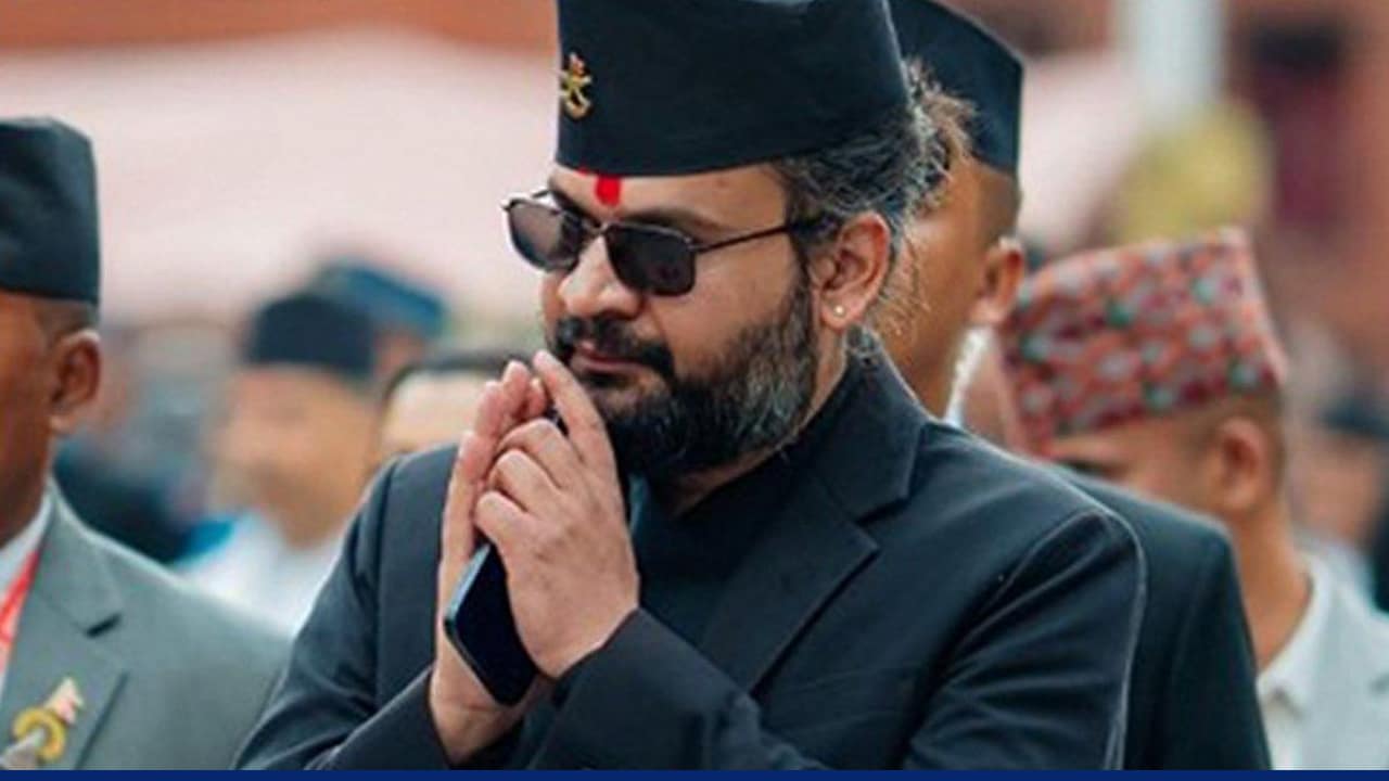 A man wearing a black traditional outfit and a Nepali-style topi, with a red tika on his forehead and sunglasses, stands with hands pressed together in a respectful greeting gesture, surrounded by others in the background.