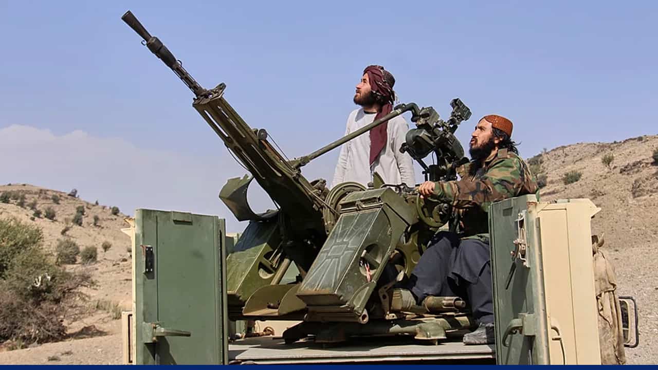 Two armed men stand on the back of a military vehicle in a desert landscape, operating a mounted anti-aircraft gun and looking upward into the sky.
