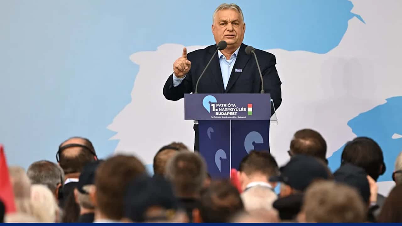 A politician speaks at a podium during a public event, gesturing with one hand while addressing an audience seated in front of him, with a backdrop featuring a stylized map.