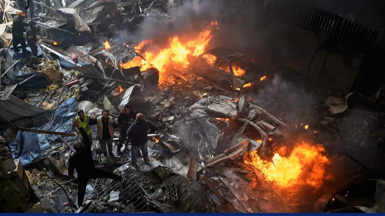 Rescue workers stand among heavy rubble and twisted metal as several cars burn with large flames and thick smoke rising in a densely damaged urban area.