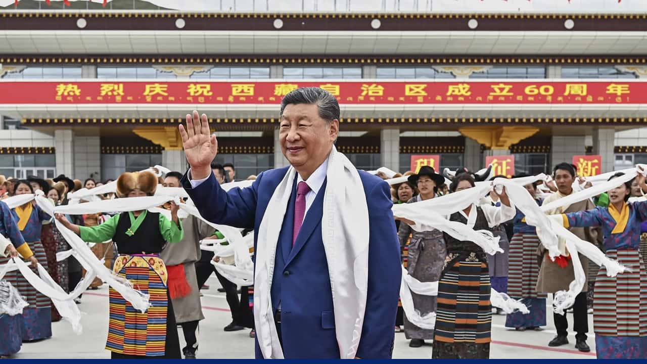 A Chinese political leader waves to a crowd while performers in traditional ethnic clothing dance with white scarves during a cultural celebration outside a large government building with a red banner.