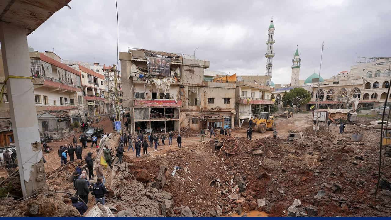 People gather around a large crater and damaged buildings in a city street following a missile strike, with a mosque visible in the background.