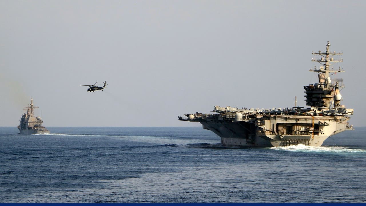 US aircraft carrier sailing at sea accompanied by a naval escort ship, with a military helicopter flying overhead.
