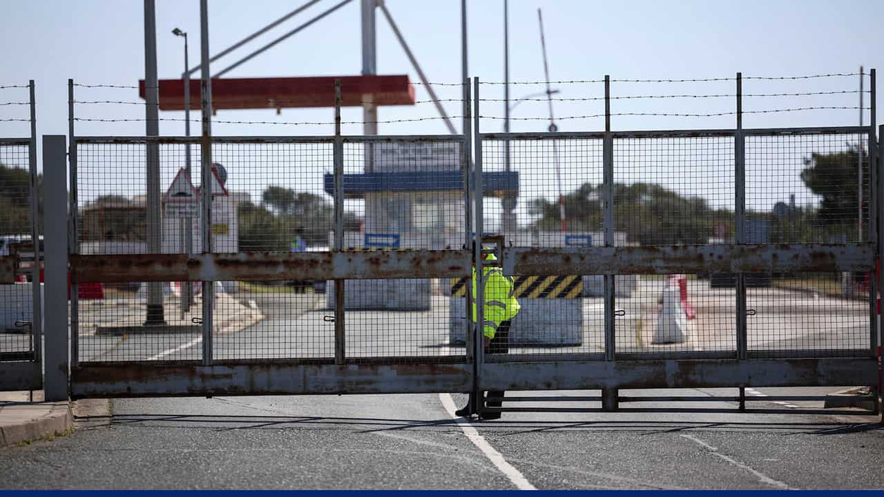 Closed security gate at a restricted area in Cyprus, with a guard in a high-visibility jacket standing behind the metal fence during heightened security measures.
