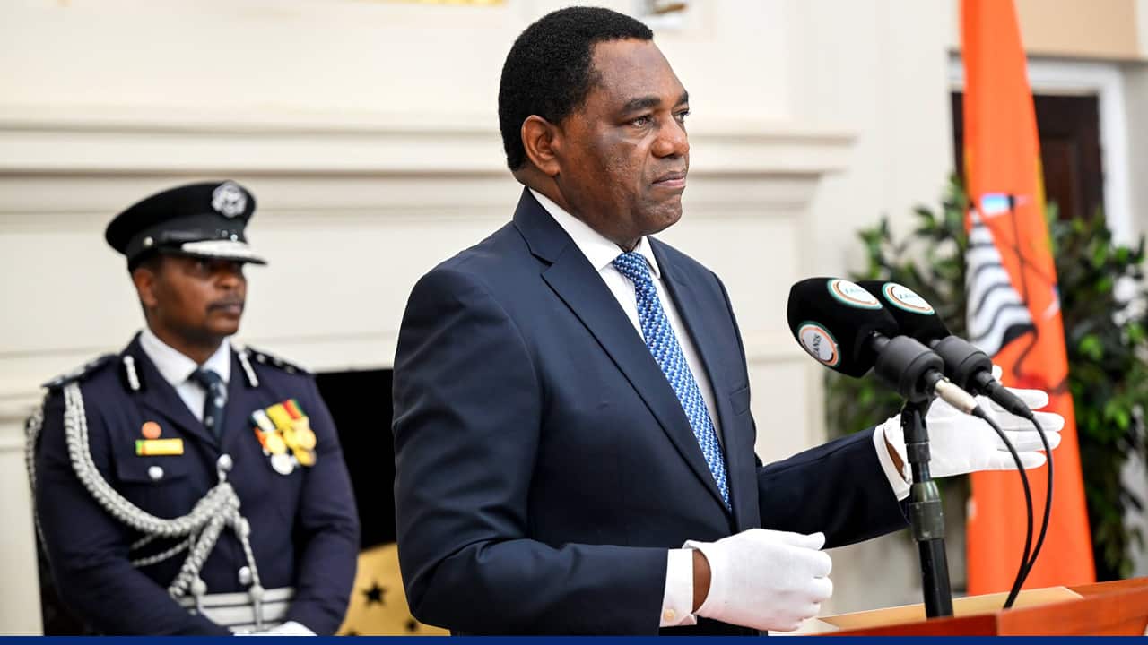 A formally dressed official in a navy suit and blue tie delivers a speech at a podium with multiple microphones, wearing white gloves, while a uniformed officer with medals stands in the background.
