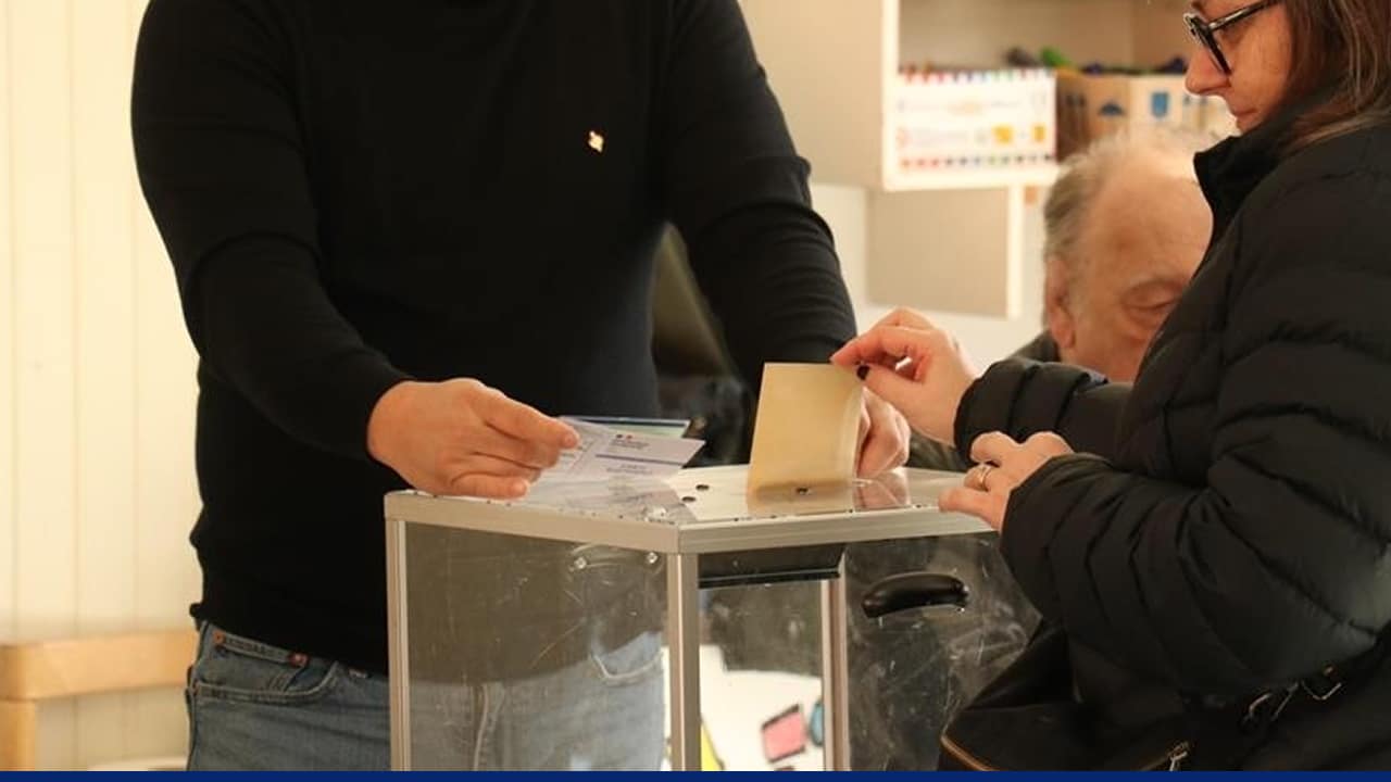Voter placing a ballot into a transparent ballot box at a polling station during local elections, with election officials assisting nearby.