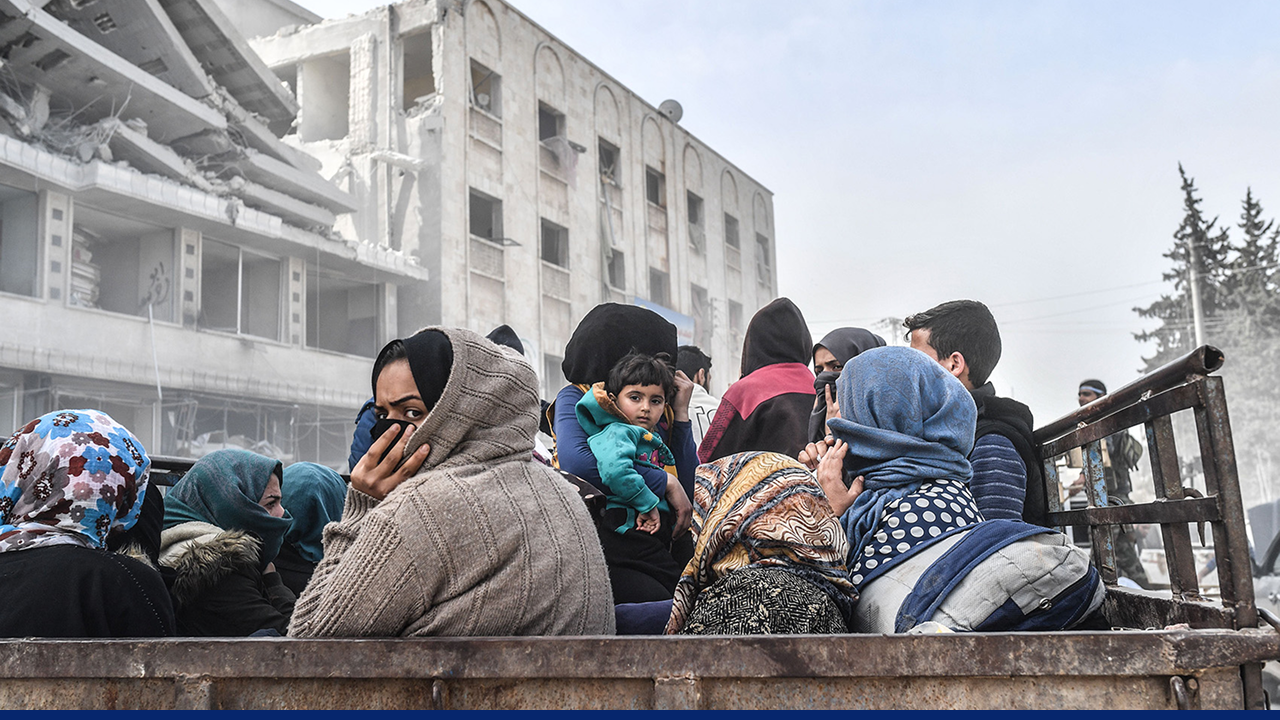 Group of Syrian civilians, including women and children, sitting in the back of a vehicle after fleeing airstrikes, with a damaged building visible in the background.